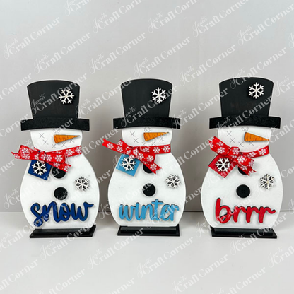 Janet's Craft Corner Snowman Shelf Sitters feature three snowmen in black hats and red or blue scarves, each with a snowflake design and festive word—ideal for winter or holiday home decor.