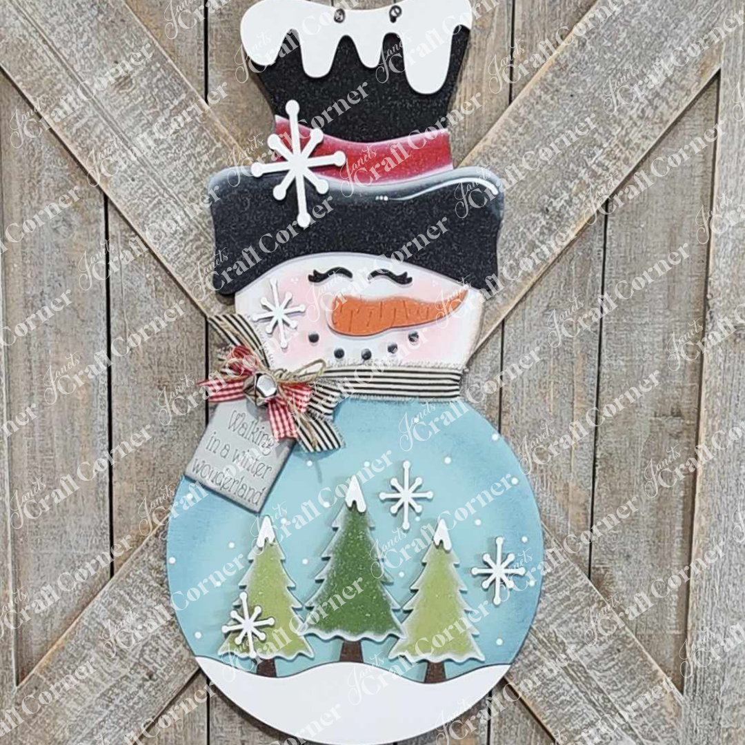 The Snowman Trees Wall/Door Hanger by Janet's Craft Corner is a DIY seasonal home decor craft kit featuring a snowman with hat, carrot nose, pink cheeks, and scarf amid green trees and snowflakes—a rustic festive touch for your space.