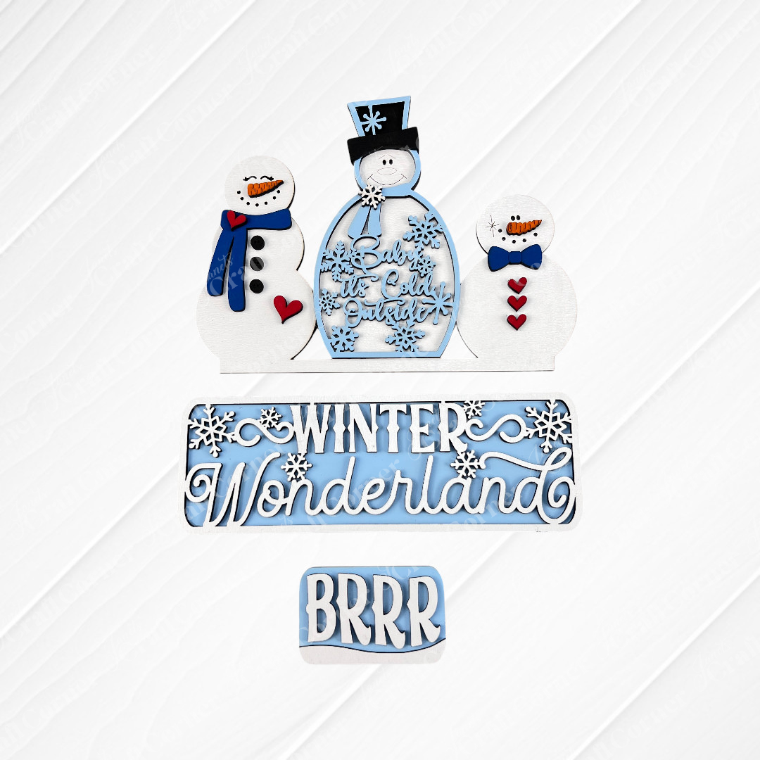 A set of hand-painted snowman cookies alongside a textured surface adorned with winter-themed signs from Janet's Craft Corner. These signs, crafted to resemble the "Winter Wonderland Interchangeable Insert - DIY Craft Kit," display festive blue and white messages like "Baby It's Cold Outside," "Winter Wonderland," and "BRRR.