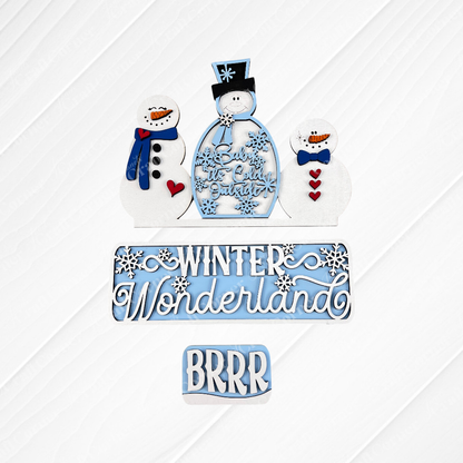 A set of hand-painted snowman cookies alongside a textured surface adorned with winter-themed signs from Janet's Craft Corner. These signs, crafted to resemble the "Winter Wonderland Interchangeable Insert - DIY Craft Kit," display festive blue and white messages like "Baby It's Cold Outside," "Winter Wonderland," and "BRRR.