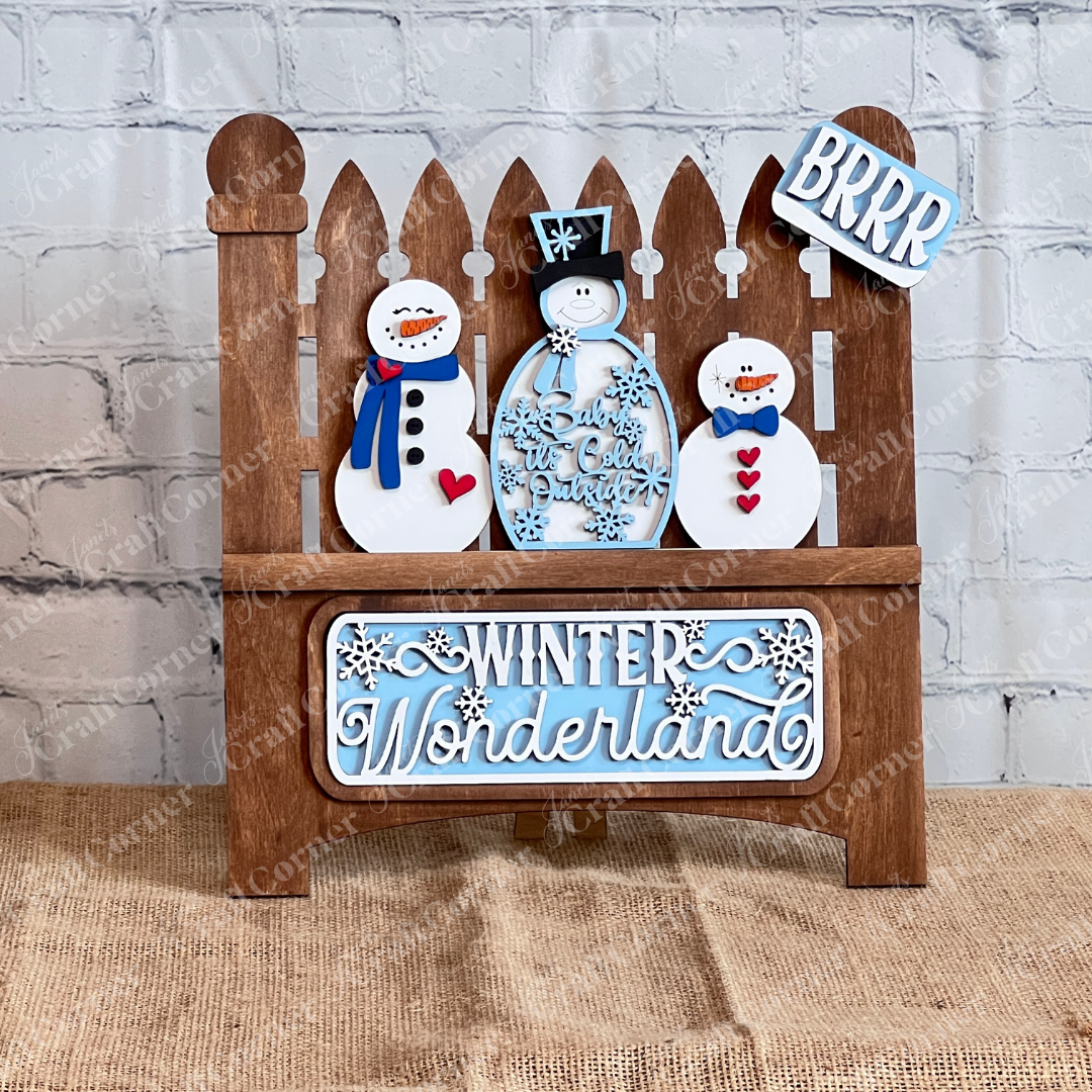 The Winter Wonderland Interchangeable Insert DIY Craft Kit by Janet's Craft Corner features a charming decorative piece depicting a garden fence with three smiling snowmen. The area below the snowmen is adorned with the words "Winter Wonderland" and on the top of the right post is "BRRR" in white and blue, all  against a white brick wall and placed on a burlap surface. It's perfect for interchangeable home decor!