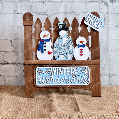 The Winter Wonderland Interchangeable Insert DIY Craft Kit by Janet's Craft Corner features a charming decorative piece depicting a garden fence with three smiling snowmen. The area below the snowmen is adorned with the words "Winter Wonderland" and on the top of the right post is "BRRR" in white and blue, all  against a white brick wall and placed on a burlap surface. It's perfect for interchangeable home decor!