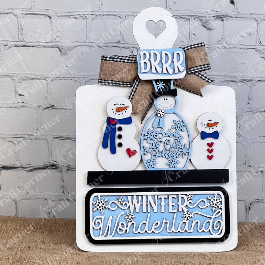 The Winter Wonderland Interchangeable Insert - DIY Craft Kit, presented by Janet's Craft Corner, allows you to craft a charming winter display featuring snowmen, hearts, and snowflakes. The scene is enhanced with phrases like "BRRR" and "Winter Wonderland" against a white brick wall backdrop on a wooden surface. This set is ideal for fans of interchangeable home decor.