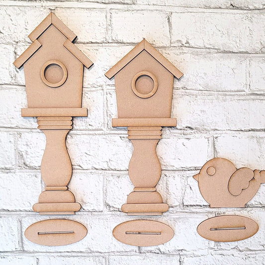 Displayed against a white brick wall, the Spindle Birdhouses Shelf Sitter by Janet's Craft Corner includes two birdhouse cutouts and a bird cutout on pedestals. This DIY kit features slots for interlocking, offering a simple, unfinished design perfect for charming shelf sitters.
