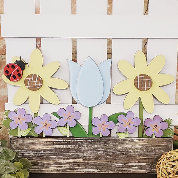 The Spring Flowers Window Box Insert from Janet's Craft Corner is a charming decorative wooden panel perfect for home décor. It features a vibrant scene with a blue tulip and yellow flowers that have button centers, all set against a backdrop of a white fence. Below, purple blooms with green leaves flourish, while a red ladybug rests on one of the spring flowers.