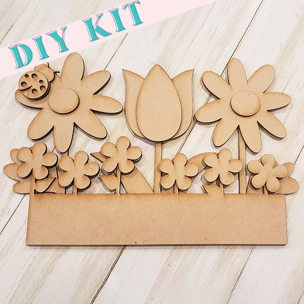 Explore the Spring Flowers Window Box Insert by Janet's Craft Corner, featuring wooden cutouts of daisies, a tulip, and small flowers with a charming ladybug on a white wooden background. Ideal for home décor, this set brings a touch of nature indoors. The phrase "DIY Kit" is elegantly displayed at the top.