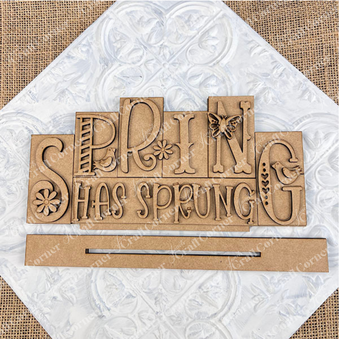 The Hello Spring Mini Word Block from Janet's Craft Corner features vibrant wooden lettering, with playful font variations. Ideal for home décor, it showcases a charming bird perched next to the letters "P" and G, a fun butterfly on the letter "N",  and delightful daisies embellishing the "S" and "I." Positioned against a white background on a burlap surface, it's perfect for adding a touch of spring to any room.