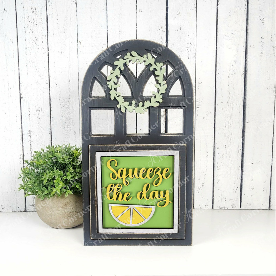 Add a pop of cheer to your space with Janet's Craft Corner Lemon Squares—a black arch with a green wreath and "Squeeze the day" sign plus lemon slice, beside a potted plant. Fun DIY or use with Swappable Squares! White wood background.