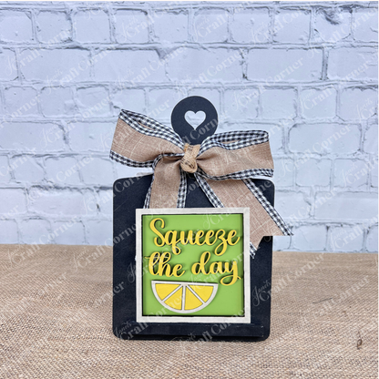 The Lemon Squares sign from Janet's Craft Corner is a small Swappable Squares piece with a bow, featuring “Squeeze the day” in yellow on green with a lemon slice. Ideal DIY seasonal decor, it's displayed on burlap before a white brick wall.