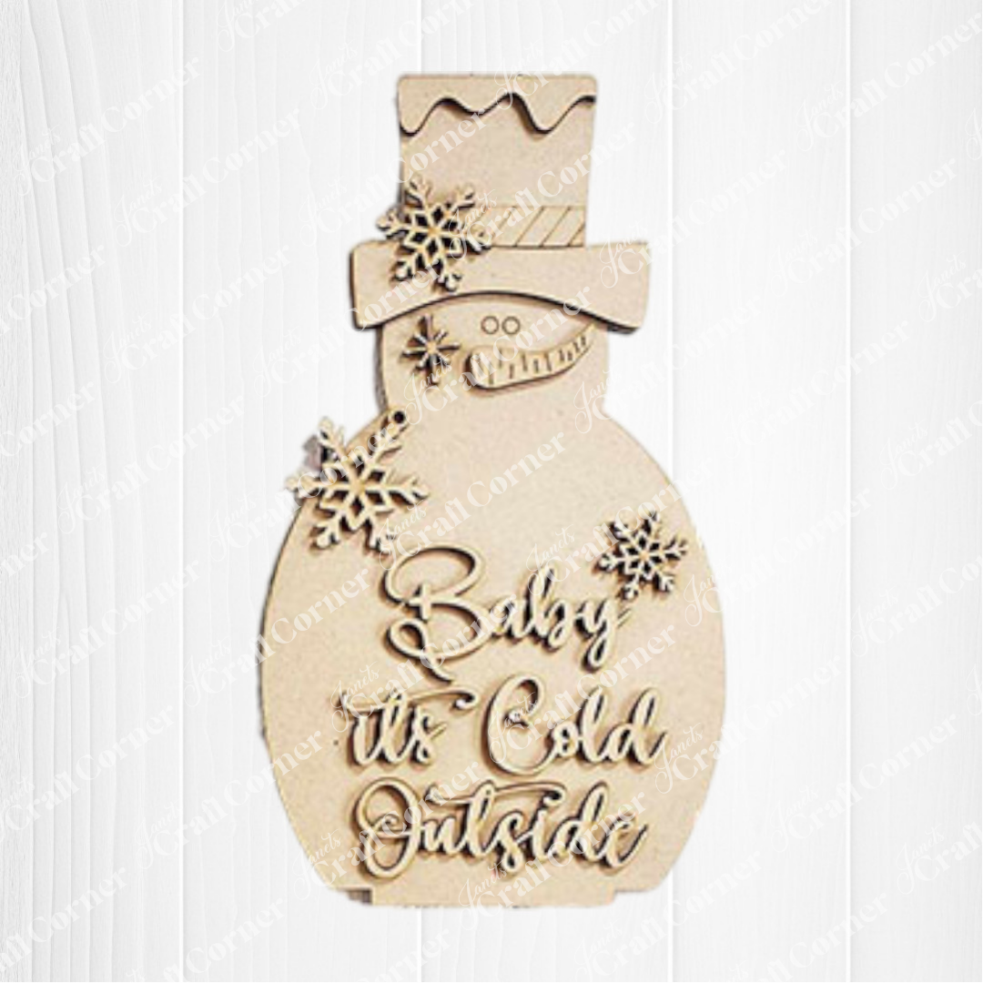 The Snowman Mini Shelf Sitters DIY craft kit by Janet's Craft Corner features a snowman cutout with snowflakes and "Baby it's cold outside" cursive text—perfect for charming holiday decor on any white wooden shelf.