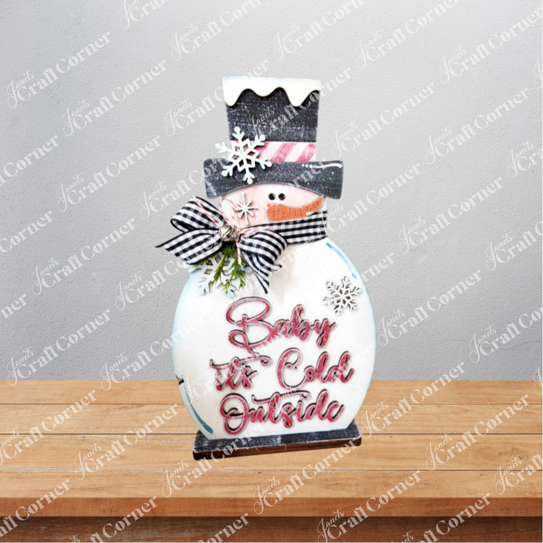 Janet's Craft Corner Snowman Mini Shelf Sitter DIY kit features a wooden snowman with a black hat, pink striped scarf, plaid bow, snowflakes, and "Baby, it's Cold Outside"—a charming holiday decor piece for any shelf. Set of 1.