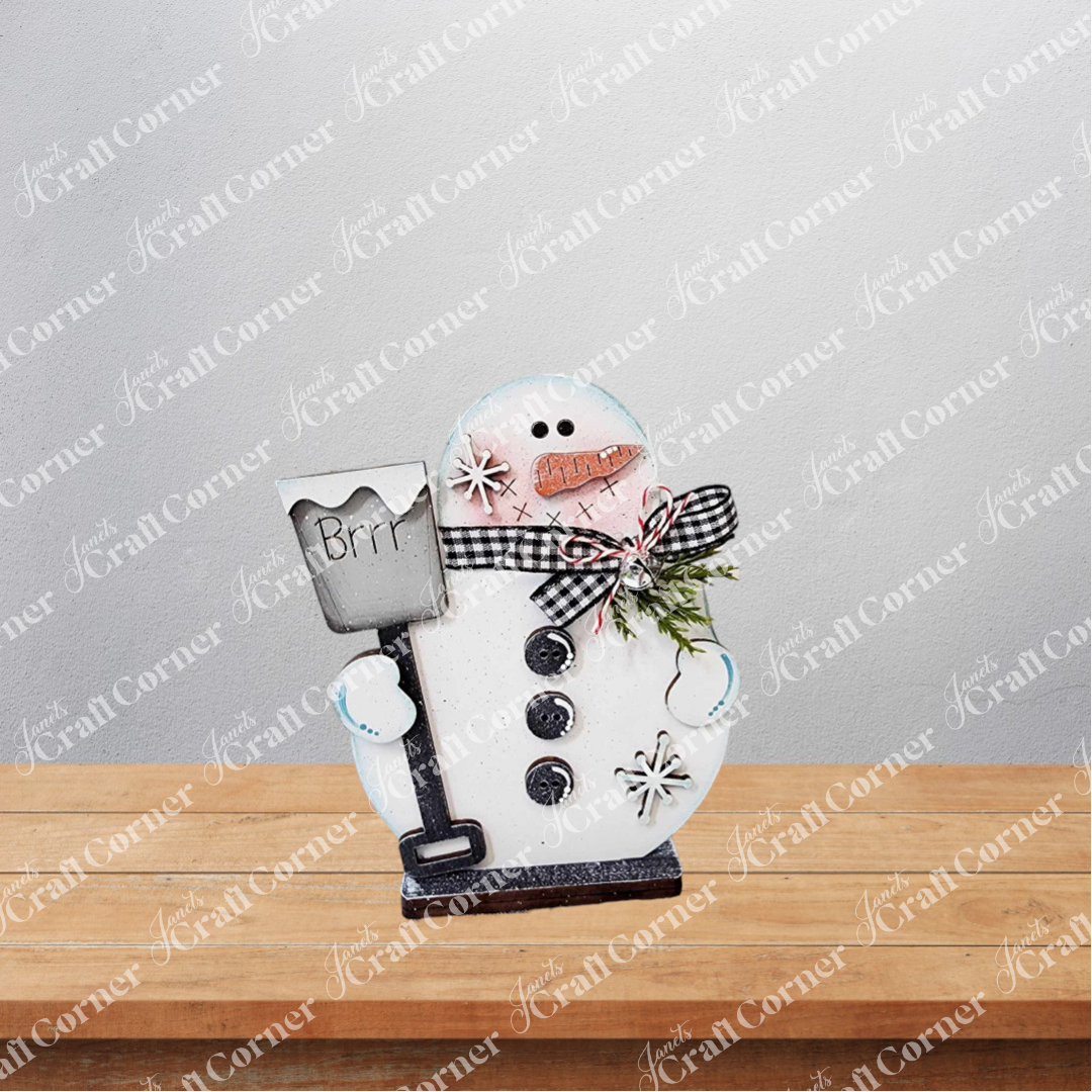 The Snowman Mini Shelf Sitter by Janet's Craft Corner is a DIY seasonal home decor craft kit featuring a wooden snowman with button details, snowflake accents, greenery, and a "Brrr" shovel—perfect for adding holiday charm to your shelf.