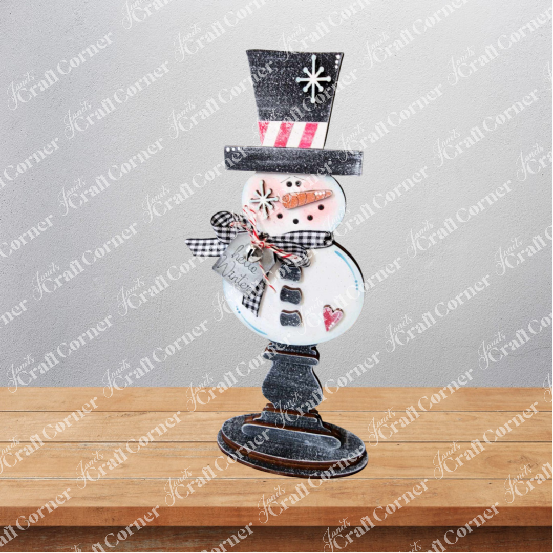 The Snowman Mini Shelf Sitter DIY craft kit by Janet's Craft Corner features a set of 1 snowman with a black top hat, pink cheeks, gingham scarf, and heart—perfect for adding whimsical charm to your holiday decor.
