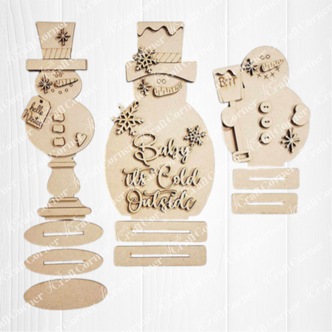 Janet's Craft Corner Snowman Mini Shelf Sitters DIY kit includes one unpainted ornament with cutout snowflakes, hats, buttons, and "Baby it's Cold Outside." Kit has disassembled pieces plus base—perfect for seasonal home decor.