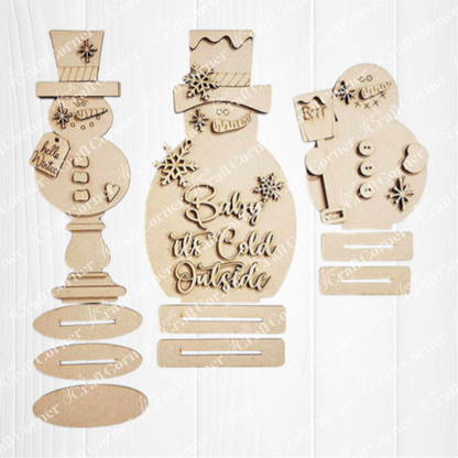 Janet's Craft Corner Snowman Mini Shelf Sitters DIY kit includes one unpainted ornament with cutout snowflakes, hats, buttons, and "Baby it's Cold Outside." Kit has disassembled pieces plus base—perfect for seasonal home decor.