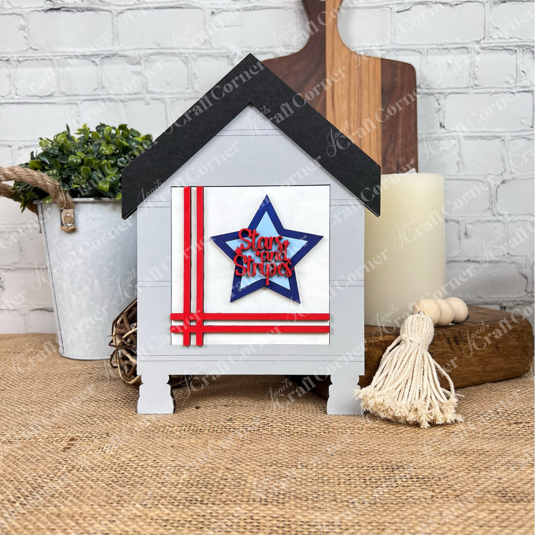 The Patriotic Squares DIY seasonal home decor craft kit from Janet's Craft Corner features a small wooden house with a black roof, red stripes, and a blue star reading "Stars and Stripes"—perfect for festive or Independence Day decorating.