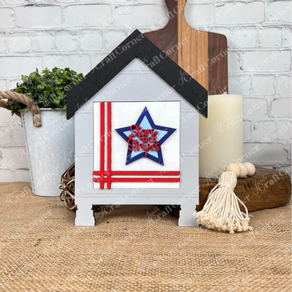 The Patriotic Squares DIY seasonal home decor craft kit from Janet's Craft Corner features a small wooden house with a black roof, red stripes, and a blue star reading "Stars and Stripes"—perfect for festive or Independence Day decorating.