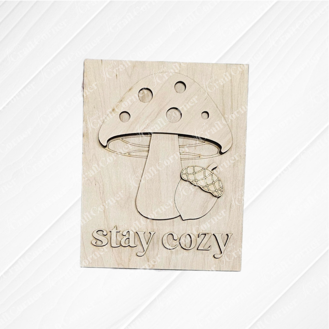 Janet's Craft Corner’s “Fall in the Forest Switchable Farmhouse Signs” DIY kit features a wooden plaque engraved with a mushroom, acorn, and “stay cozy.” Ideal for woodland or Fall farmhouse decor; light wood grain enhances its rustic charm.