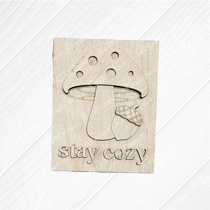 Janet's Craft Corner’s “Fall in the Forest Switchable Farmhouse Signs” DIY kit features a wooden plaque engraved with a mushroom, acorn, and “stay cozy.” Ideal for woodland or Fall farmhouse decor; light wood grain enhances its rustic charm.