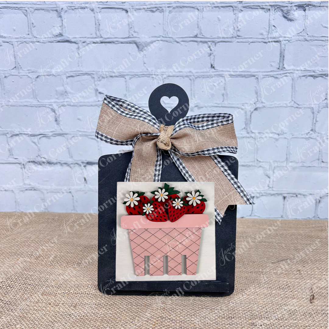A black clipboard with a heart cutout and bow displays Janet's Craft Corner's Strawberry Swappable Squares DIY seasonal home decor kit, featuring a pink flowerpot with strawberries—perfect tiered tray decor against a brick wall.
