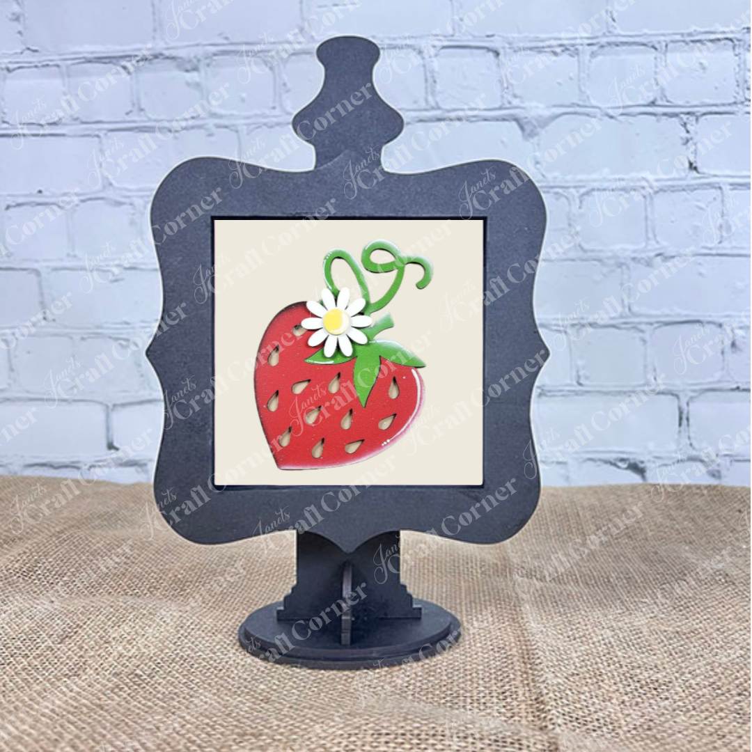 A decorative black stand displays Janet's Craft Corner’s Strawberry Swappable Squares – a DIY seasonal home decor kit with a red strawberry, green leaves, yellow seeds, and a white daisy. Shown on burlap with a white brick wall backdrop.