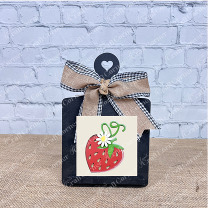 A small black chalkboard with a heart cutout and ribbon bow displays the Strawberry Swappable Squares DIY craft kit by Janet's Craft Corner—featuring a strawberry with a white flower, perfect for seasonal decor accents.