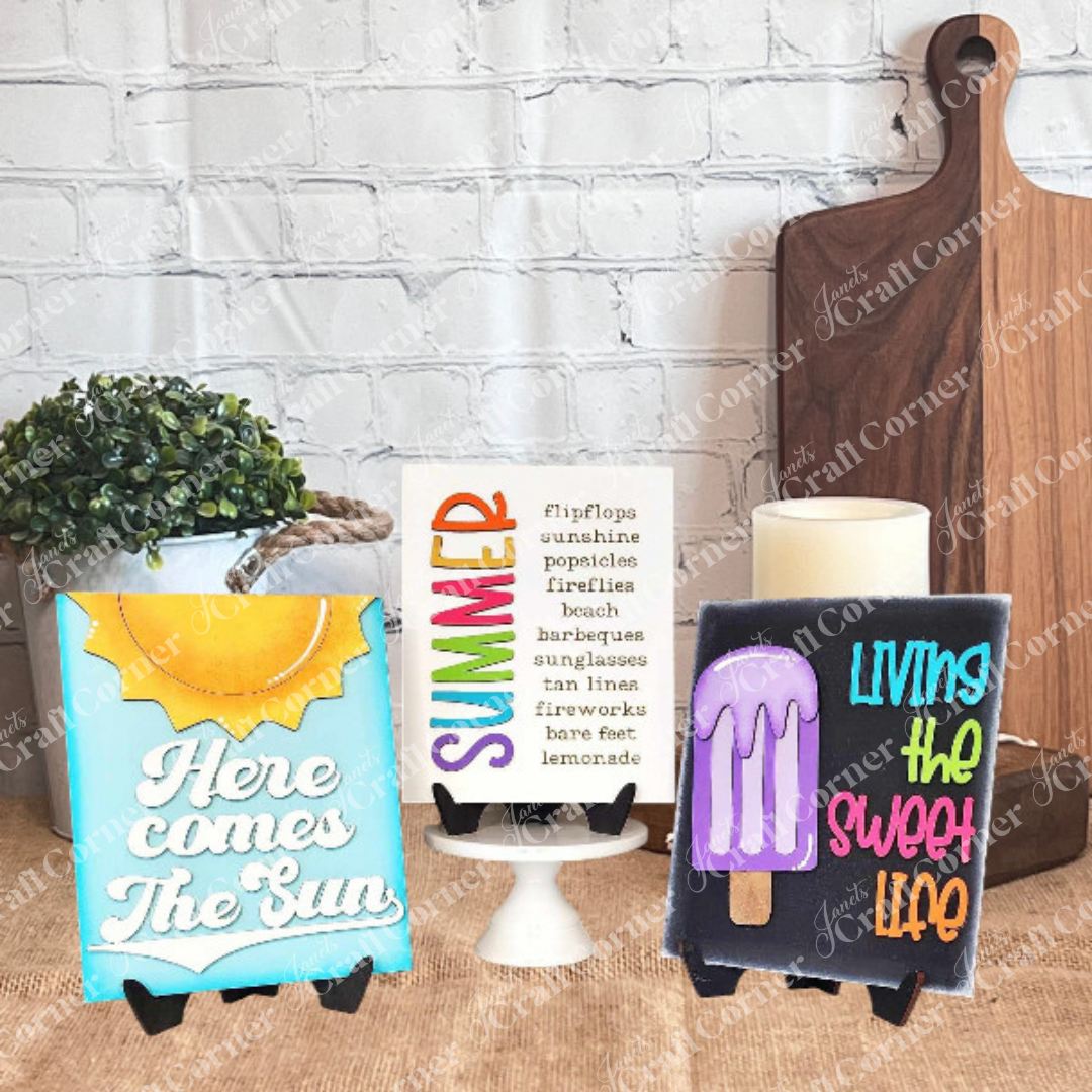 A vibrant Summer Interchangeable Farmhouse Sign from Janet’s Craft Corner is displayed on a stand, reading “Here comes the sun,” with a wooden board in the background. DIY seasonal home decor craft kit – Set of 1.