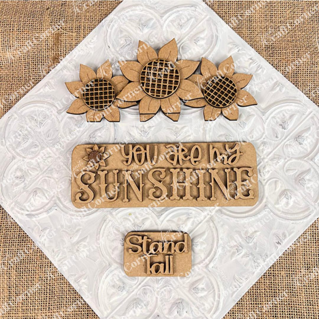 Sunflower accents on a textured backdrop: featuring three sunflowers at the top, a central plaque displaying "YOU ARE MY SUNSHINE," and a smaller plaque with "Stand Tall" positioned below. This delightful DIY kit from Janet's Craft Corner, known as the Sunflower Interchangeable Insert, is ideal for fans of versatile home decor themes.