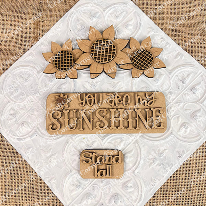 Sunflower accents on a textured backdrop: featuring three sunflowers at the top, a central plaque displaying "YOU ARE MY SUNSHINE," and a smaller plaque with "Stand Tall" positioned below. This delightful DIY kit from Janet's Craft Corner, known as the Sunflower Interchangeable Insert, is ideal for fans of versatile home decor themes.