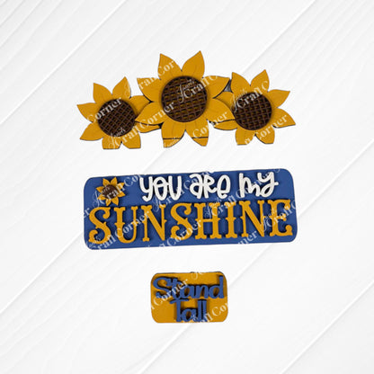 In the Sunflower Interchangeable Insert DIY home decor kit by Janet's Craft Corner, decorative cookies are beautifully set against a white embossed background, embodying the sunflower theme. Among these, three cookies are crafted into sunflower shapes; one is inscribed with "You are my sunshine" alongside a charming sunflower graphic, while another smaller one bears the message "Stand tall." The color palette features vibrant yellow, soothing blue, and earthy brown tones.