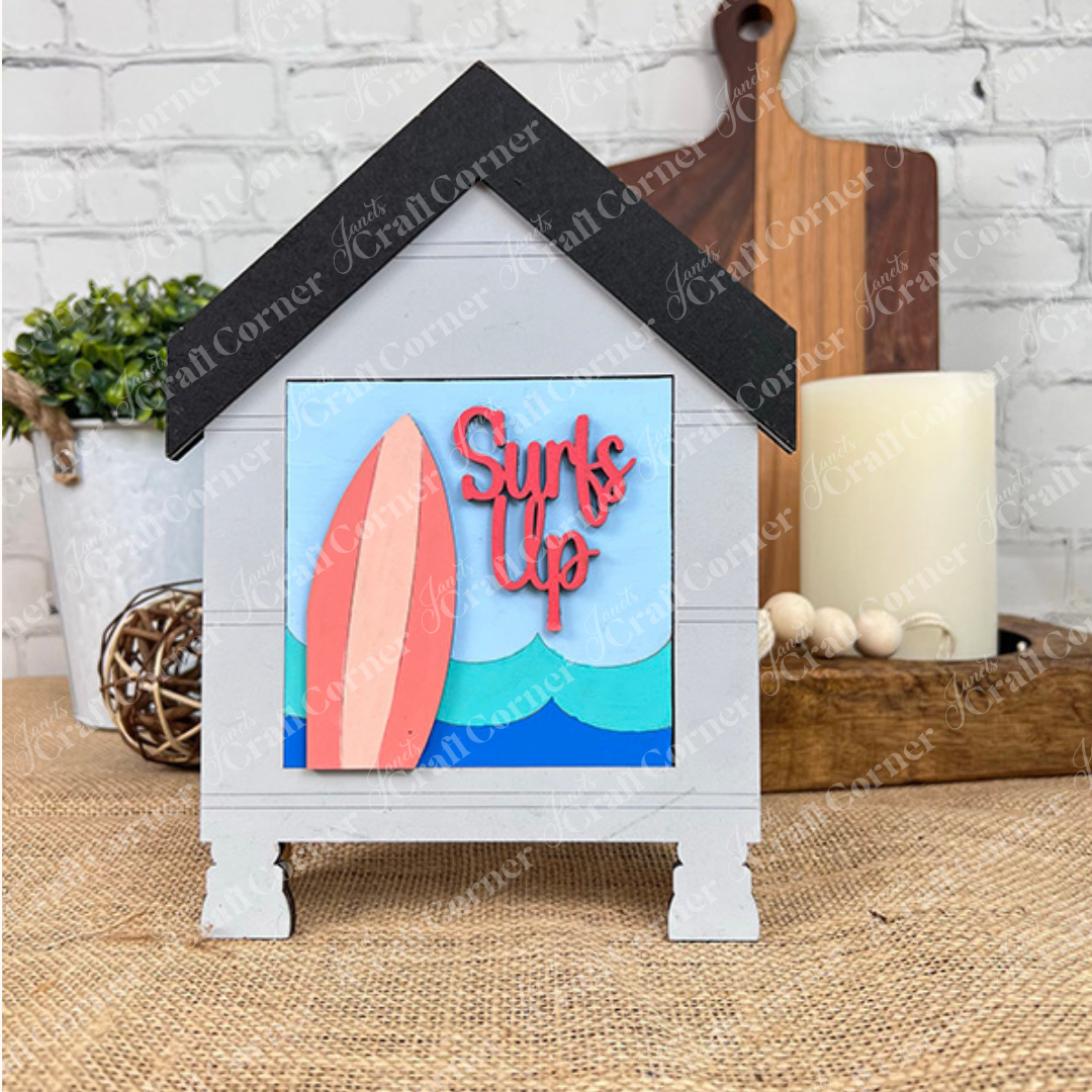 The Summer Beach Squares DIY seasonal home decor craft kit by Janet's Craft Corner features a decorative surf shack sign with a black roof, pink surfboard, and "Surf's Up" in red on blue—perfect for adding summer vibes to your space.