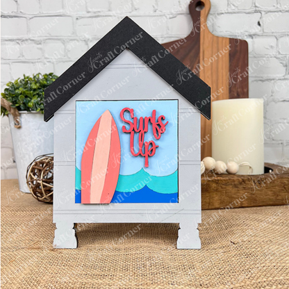 The Summer Beach Squares DIY seasonal home decor craft kit by Janet's Craft Corner features a decorative surf shack sign with a black roof, pink surfboard, and "Surf's Up" in red on blue—perfect for adding summer vibes to your space.