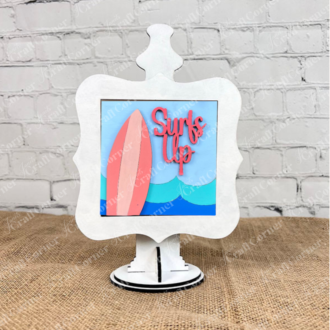 The Summer Beach Squares DIY seasonal home decor craft kit from Janet's Craft Corner features a white decorative frame on burlap with a colorful "Surf's Up" sign showing a pink surfboard and blue wave, perfect for summer displays.