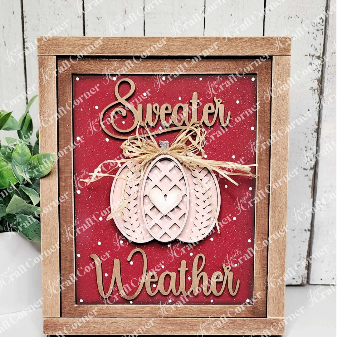 Introducing the "Sweater Weather Interchangeable Sign" by Janet's Craft Corner, a DIY home decor craft kit. This wooden-framed sign features "Sweater Weather" in elegant cursive on a vibrant red background, adorned with a decorative pumpkin topped with a bow at the center. Enhance your space effortlessly with this versatile decor piece, complementing any setting alongside a small potted plant against a white wooden wall backdrop.
