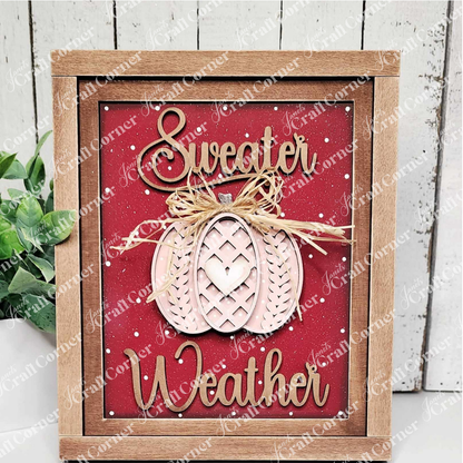 Introducing the "Sweater Weather Interchangeable Sign" by Janet's Craft Corner, a DIY home decor craft kit. This wooden-framed sign features "Sweater Weather" in elegant cursive on a vibrant red background, adorned with a decorative pumpkin topped with a bow at the center. Enhance your space effortlessly with this versatile decor piece, complementing any setting alongside a small potted plant against a white wooden wall backdrop.