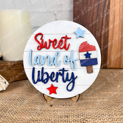 The Patriotic Mini Shelf Sitters DIY Craft Kit by Janet's Craft Corner features a round wooden sign with "Sweet land of liberty" in red and blue, stars, and a popsicle, displayed on an easel atop burlap for Americana décor.