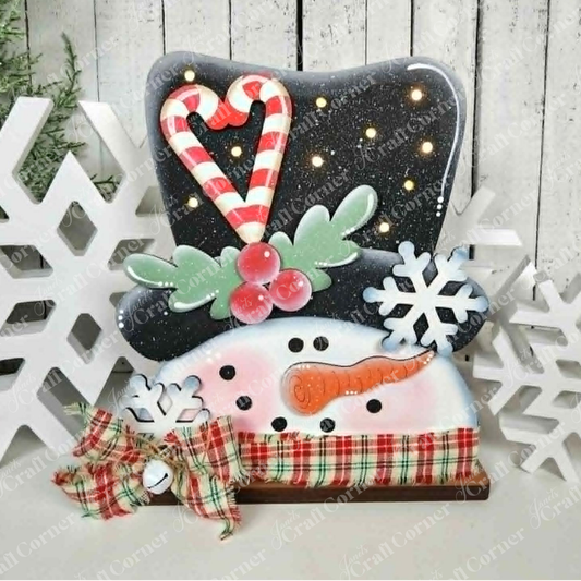 Brighten up your holiday décor with the Sweet Snowman Lighted Shelf Sitter from Janet’s Craft Corner. This DIY home decor craft kit features a cheerful snowman face adorned with a carrot nose, peppermint candy cane, holly leaves, plaid ribbon, and delicate snowflakes. Displayed on a decorative stand with a wintry background and white snowflake accents, this charming shelf sitter set is perfect for adding festive flair to your seasonal display or as part of your holiday crafting projects. Set of 1.