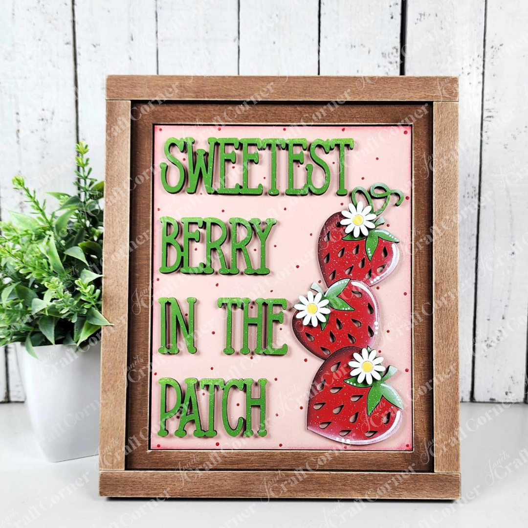 A painted sample of a sign from Janet's Craft Corner's Sweetest Berry Switchable Farmhouse Signs DIY home decor craft kit is displayed against a white shiplap wall. The sign features wood cut-outs reading "Sweetest Berry in the Patch" in green with red strawberry accents and white flowers on a pink background—perfect for seasonal home decor.