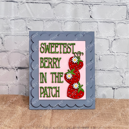 Janet's Craft Corner Single Scallop Trim Farmhouse Frame features "Sweetest Berry In The Patch" in green, with two strawberries and white daisies on a pink background, propped against a white brick wall.