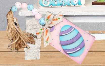 A handcrafted piece from Janet’s Craft Corner’s Easter Bunny Decor Set - Painted, featuring a blue striped egg tag with carrot-patterned ribbon, pastel beads, and a raffia tassel, rests on a wooden surface in front of a white shelf—an ideal farmhouse-style accent for your DIY home decor kit.
