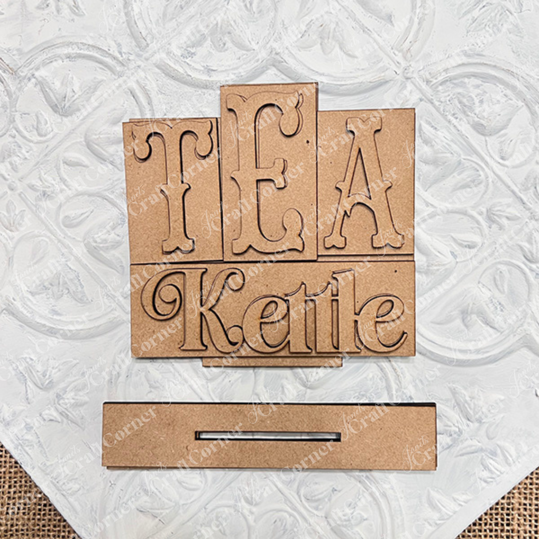 The Tea Kettle Mini Word Block Shelf Sitter from Janet's Craft Corner is a DIY decor kit. It features "TEA Kettle" in decorative font on a background of wooden blocks, with an unfinished look and a slot for easy crafting, perfect for tea lovers.