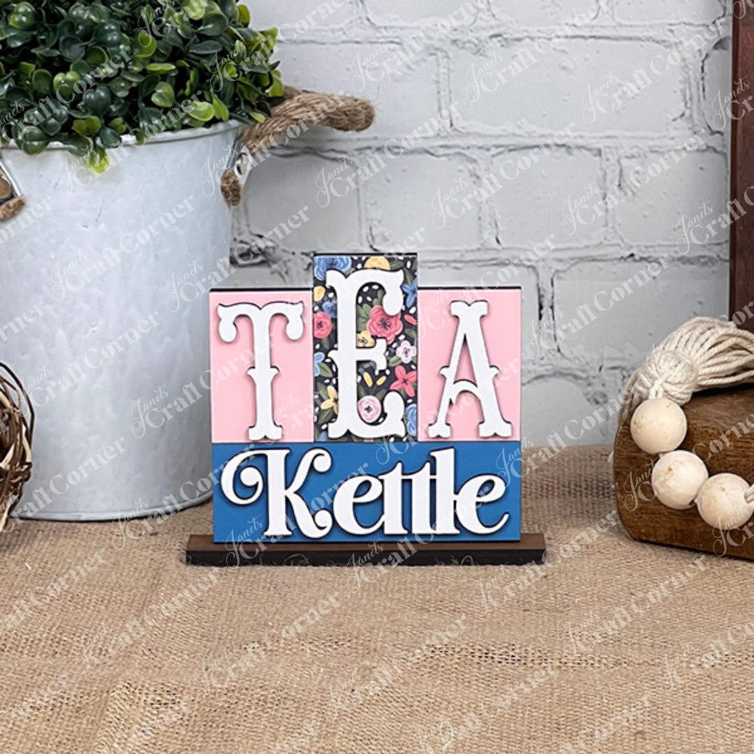 The Tea Kettle Mini Word Block from Janet's Craft Corner features the word "TEA" prominently against a floral background, with "Kettle" below. This delightful piece, ideal for home décor enthusiasts or as a DIY kit project, is displayed on a burlap surface surrounded by plants and rustic elements.