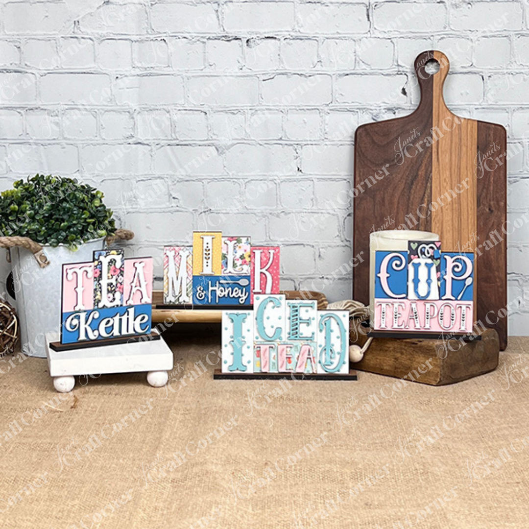 Painted samples of the tea mini word block wood cut-out DIY craft kits by Janet's Craft Corner. 4 signs shown including tea kettle, milk & honey, iced tea and cup teapot. The samples are painted in shades of blue & pink with some scrapbook paper backgrounds behind the letters.