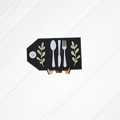 The "Thankful Mini Shelf Sitters" DIY craft kit by Janet's Craft Corner features a small black board with fork, knife, and spoon illustrations and green leaf accents—perfect for creating unique seasonal home decor.