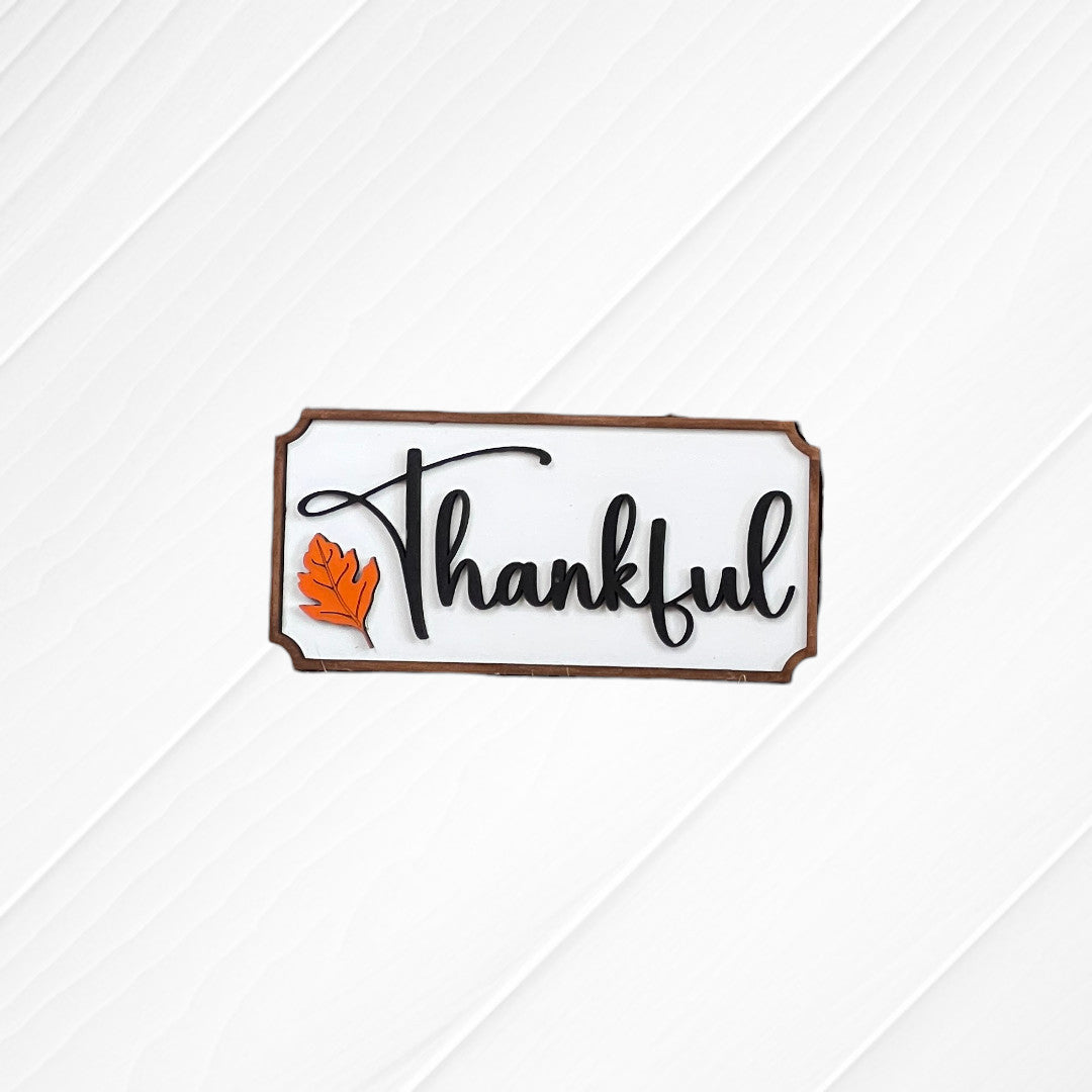 The Thankful Mini Shelf Sitter from Janet's Craft Corner is a DIY seasonal home decor kit featuring "Thankful" in black cursive and an orange leaf on a white wood background—ideal for personalized autumn decorations.