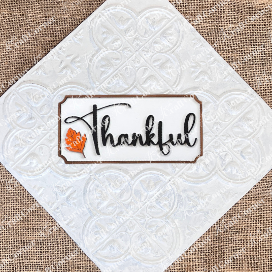 The Thankful Mini Shelf Sitter from Janet's Craft Corner is a DIY seasonal home decor kit featuring "Thankful" in black cursive and an orange leaf on a white wood background—ideal for personalized autumn decorations.