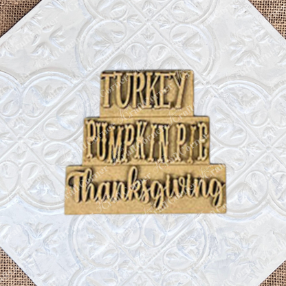 The Give Thanks/Thanksgiving Tiered Tray Kit by Janet's Craft Corner includes a stacked wooden sign featuring the words "Turkey," "Pumpkin Pie," and "Thanksgiving" arranged in three tiers, ideal for decorating your tiered tray. The kit also contains two separate wooden pieces to add delightful accents to your home décor.