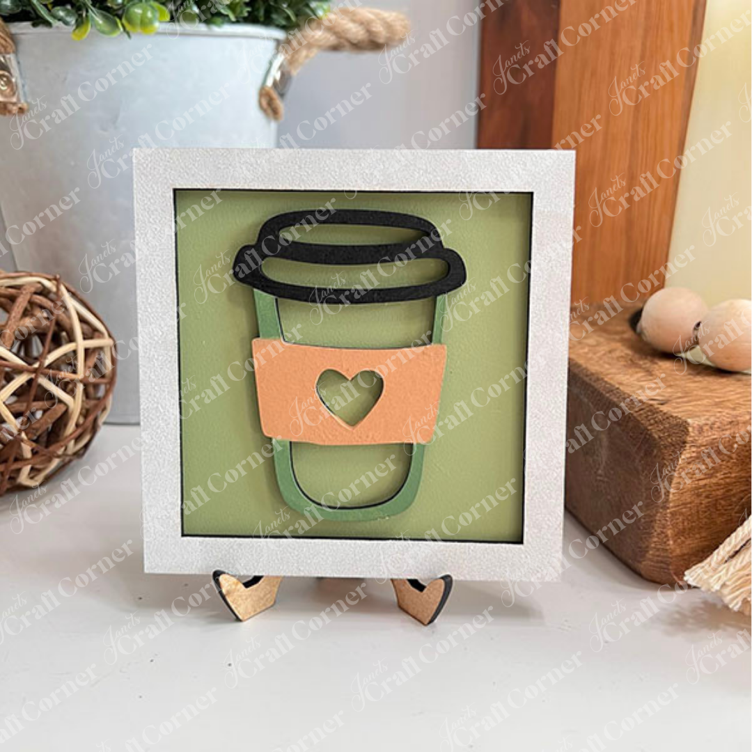 Coffee Bar Swappable Squares by Janet's Craft Corner: a framed drawing of a coffee cup with a coral sleeve and green heart, displayed on wood risers as charming DIY home decor against a white brick wall.