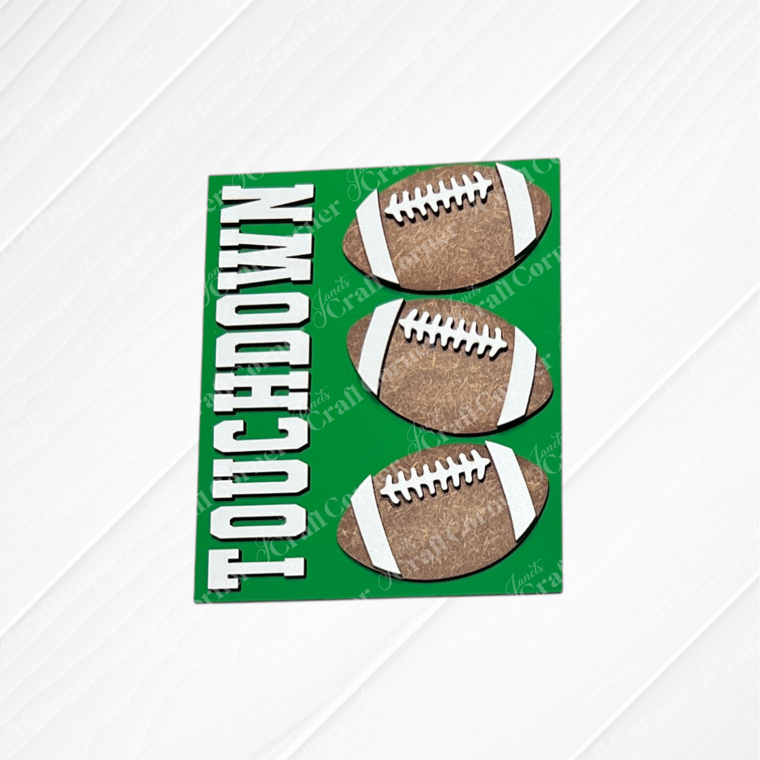 Introducing Football Farmhouse Signs by Janet’s Craft Corner—a vibrant green card featuring three brown footballs and the bold white “TOUCHDOWN” lettering along the left side. The wood-textured white background adds rustic charm, making this piece a perfect addition to your switchable farmhouse sign or interchangeable seasonal display from any DIY home decor kit.