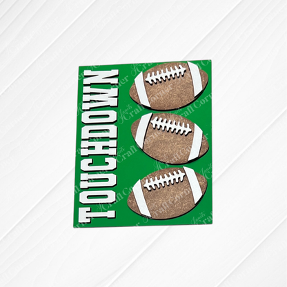 Introducing Football Farmhouse Signs by Janet’s Craft Corner—a vibrant green card featuring three brown footballs and the bold white “TOUCHDOWN” lettering along the left side. The wood-textured white background adds rustic charm, making this piece a perfect addition to your switchable farmhouse sign or interchangeable seasonal display from any DIY home decor kit.